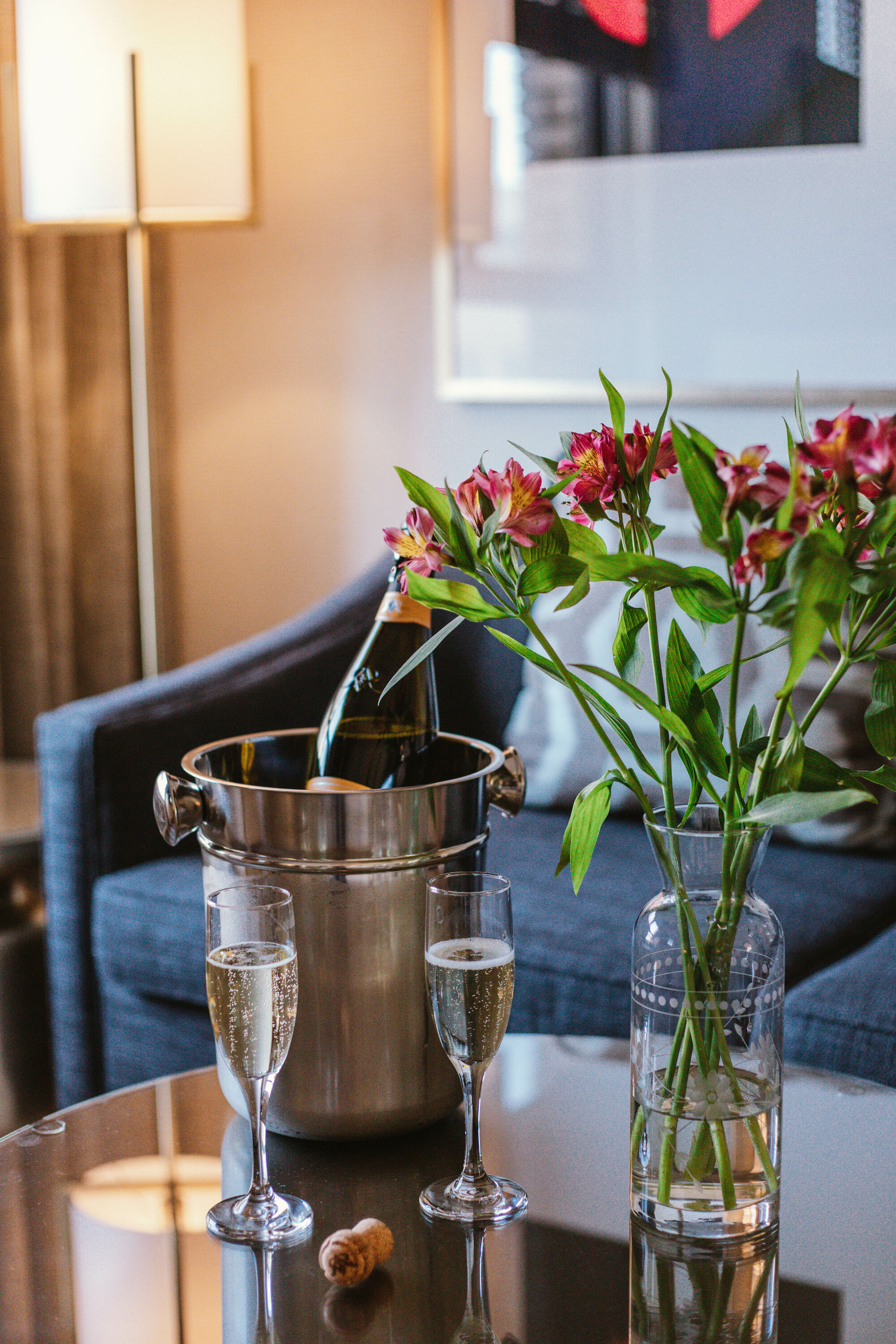 A bottle of champagne chilling in an ice bucket, two flutes ready, a vase of pink flowers, and a glass of water on a coffee table.