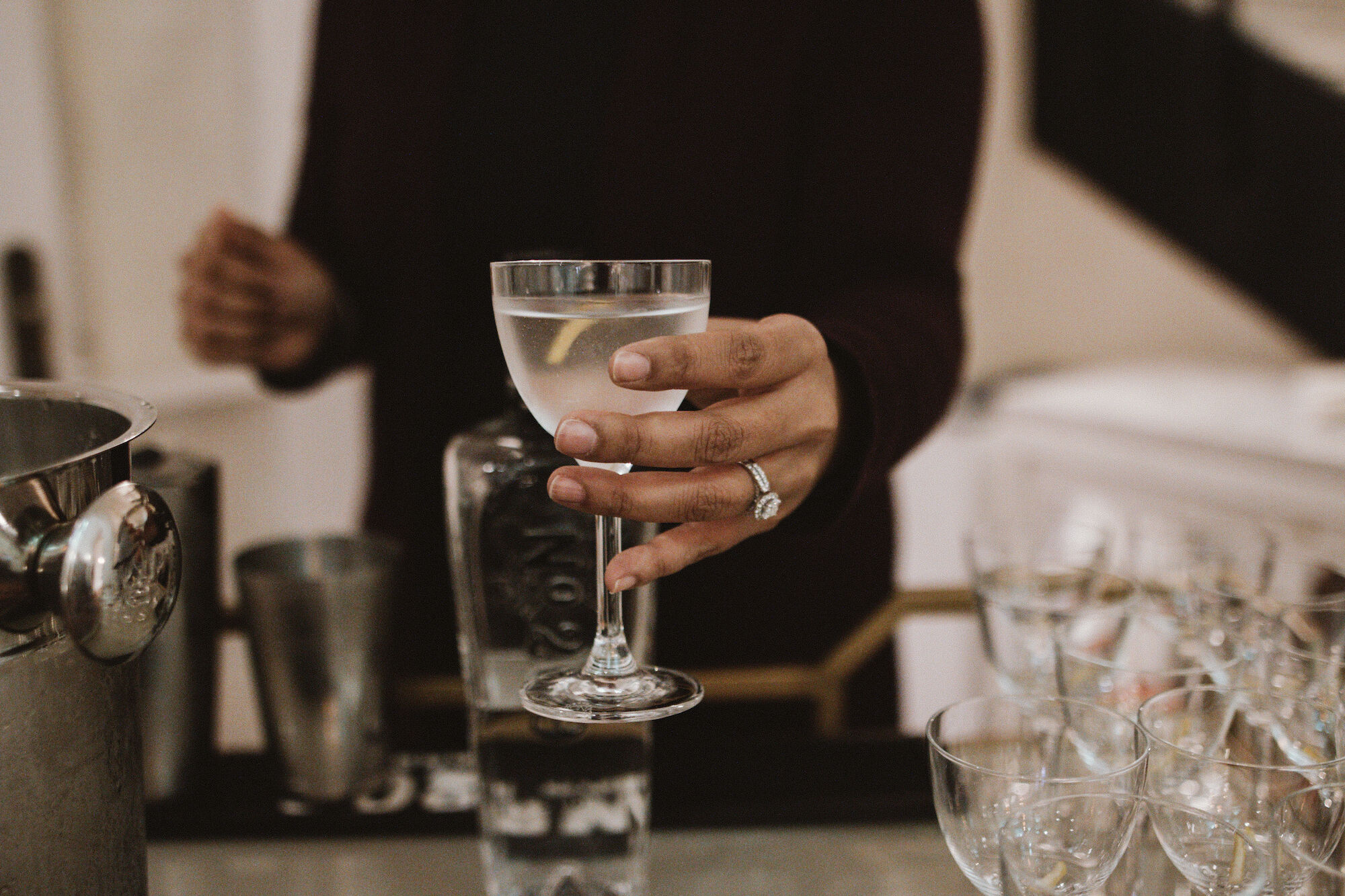 A person holding a cocktail glass, with additional glasses and bar tools on a counter, suggesting a bar or party setting.