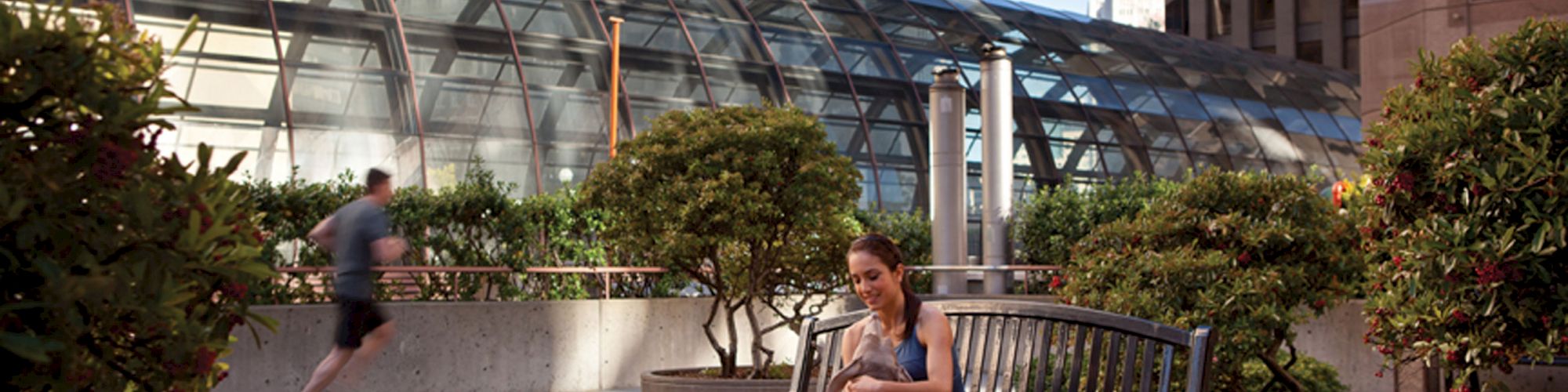 A woman is sitting on a bench in an urban garden setting with trees and buildings, and a person is jogging in the background.