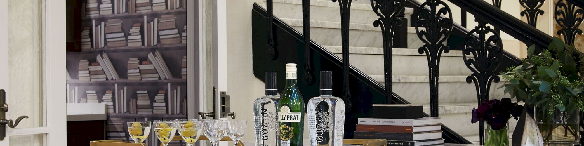 A bar cart with bottles and glassware stands in front of a staircase with ornate railings; a bookshelf wallpaper is in the background.