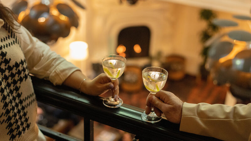 Two people holding drinks on a balcony overlooking a room with a fireplace