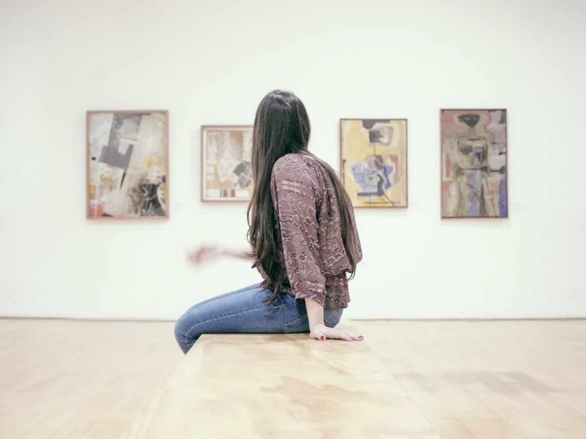 A person with long hair sits on a bench in an art gallery, facing four abstract paintings hung on a white wall.