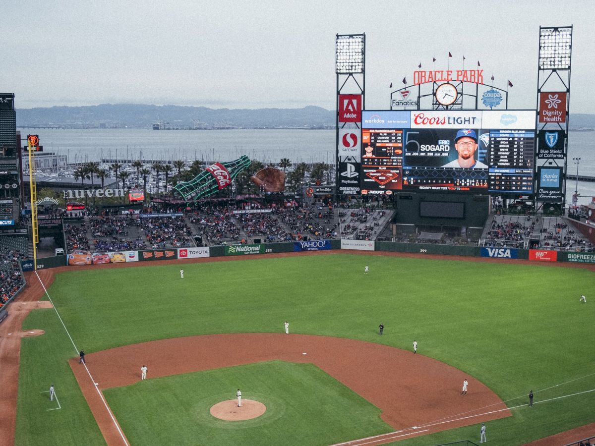 A baseball stadium with a game in progress, spectators in the stands, and a waterfront backdrop beyond the outfield.