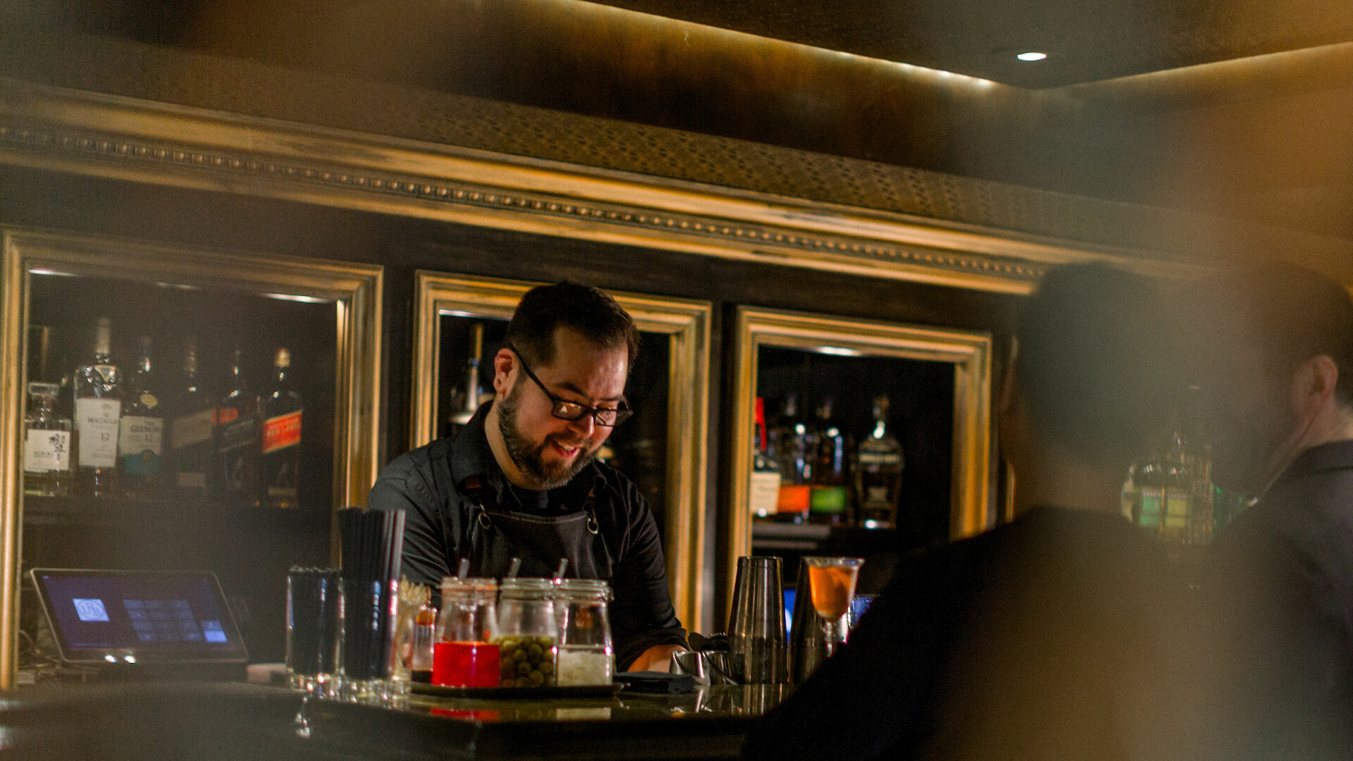 A bartender is smiling and preparing drinks behind a bar, with patrons seated at the counter in a dimly lit atmosphere.