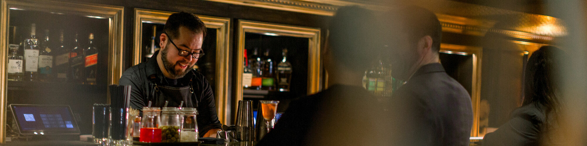 A bartender is smiling and preparing drinks behind a bar, with patrons seated at the counter in a dimly lit atmosphere.