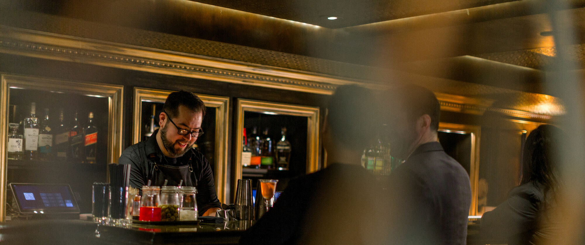 A bartender is smiling and preparing drinks behind a bar, with patrons seated at the counter in a dimly lit atmosphere.