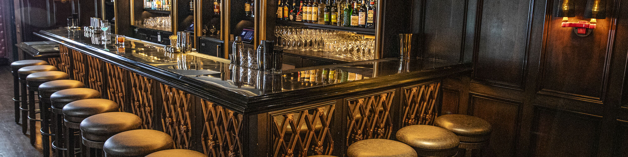 The image shows an empty bar with wooden stools and a long counter, featuring shelves stocked with various bottles and dim lighting for ambiance.