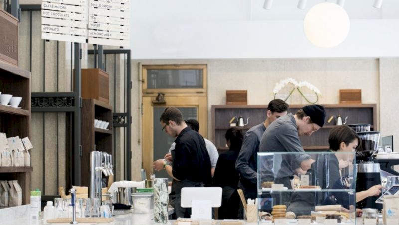 A bustling coffee shop with people behind the counter and shelves with various items.