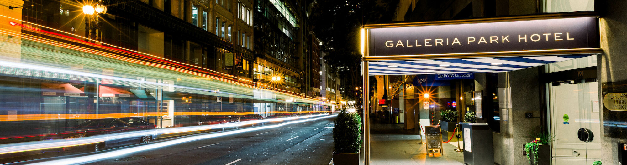 Night street scene with light trails from moving vehicles and a view of the Galleria Park Hotel entrance, with some streetlights glowing.