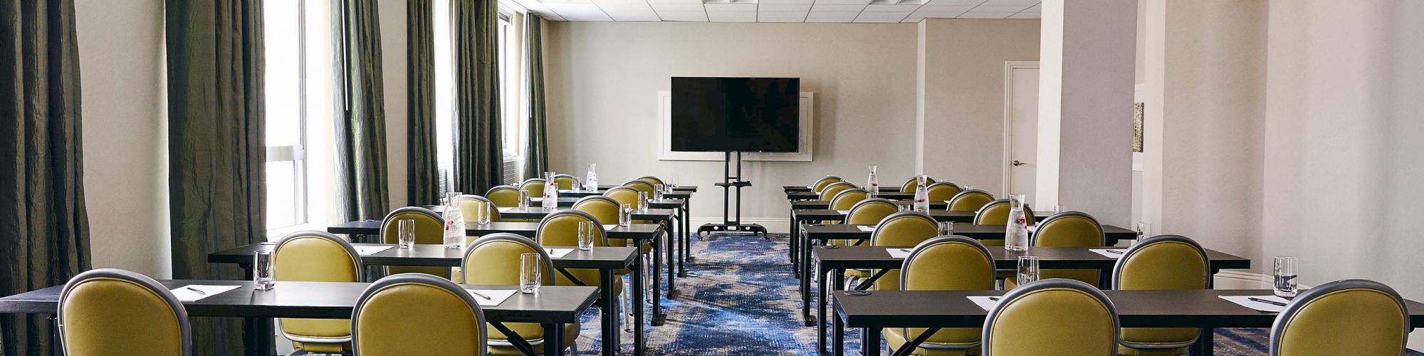 The image shows an empty conference room with rows of chairs and tables facing a TV screen, all set on a blue patterned carpet.