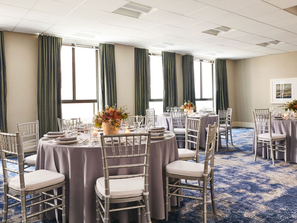 The image shows a well-lit banquet hall with round tables covered in grey tablecloths, adorned with floral centerpieces, and surrounded by white chairs.