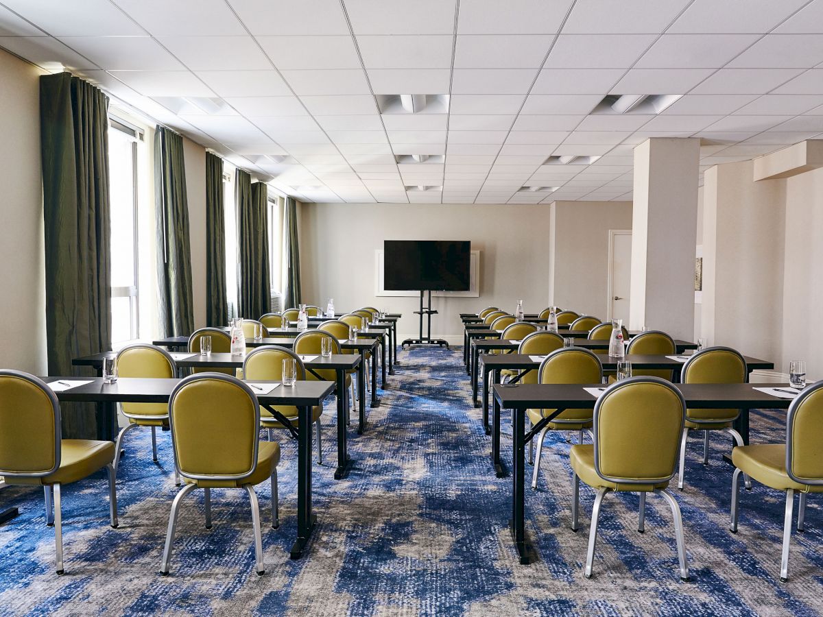 A conference room with rows of green chairs and tables facing a TV screen, featuring blue carpet and large windows with green curtains.