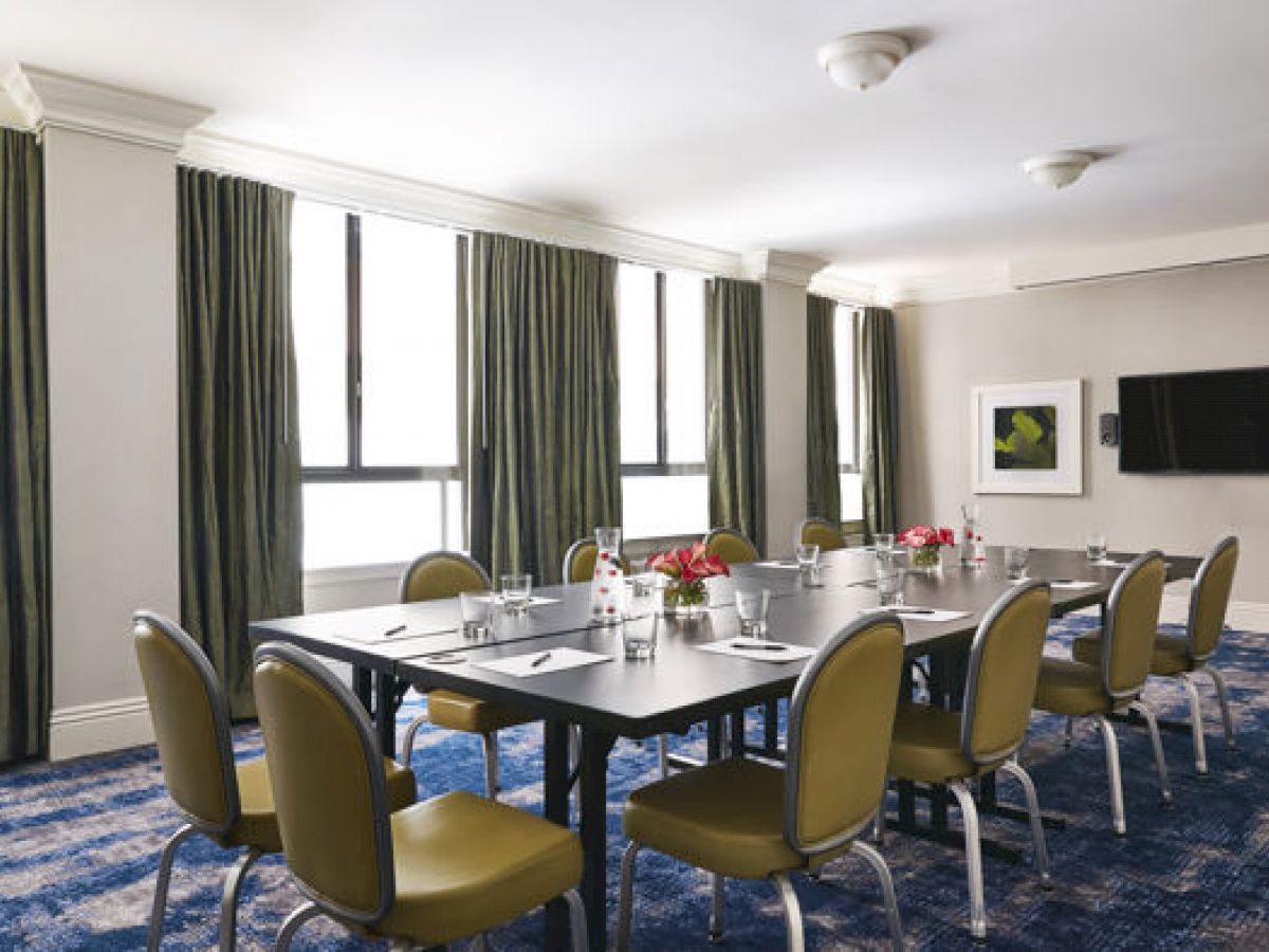 A conference room with a large table, green chairs, a TV on the wall, windows with dark curtains, and a carpeted floor.