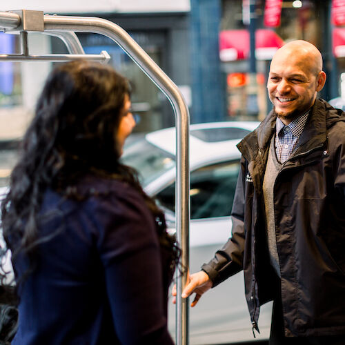 A person greets a woman with a smile outdoors, next to a car and a luggage cart, with buildings and a plant visible in the background.