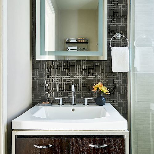 A modern bathroom features a sink with an under-cabinet, a decorative mirror, tiled backsplash, towel ring, and a flower vase.