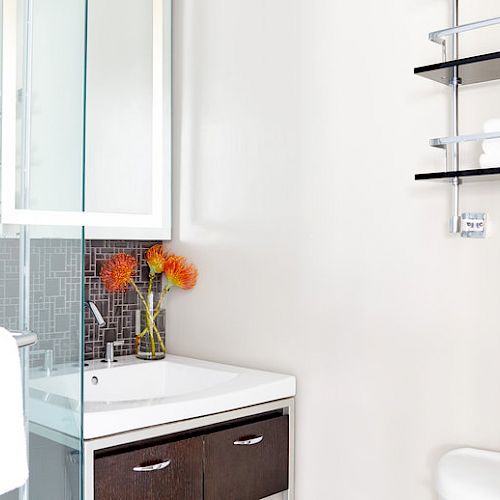 This image shows a modern bathroom with a sink, cabinets, a glass shower, shelves with towels and toiletries, and a couple of orange flowers in a vase.