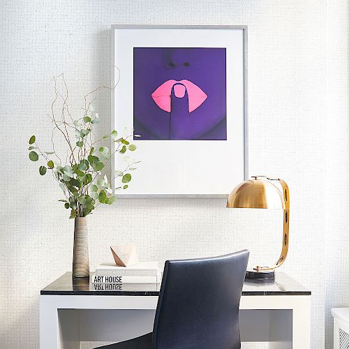 A modern desk setup with a chair, lamp, potted plant, books, and a framed artwork featuring a pink umbrella on a purple background hanging above the desk.