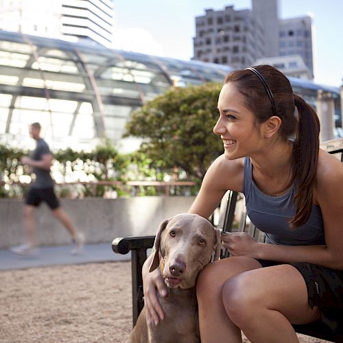 A woman sitting on a bench with a dog, smiling, in an urban park area with a man jogging in the background and buildings around.