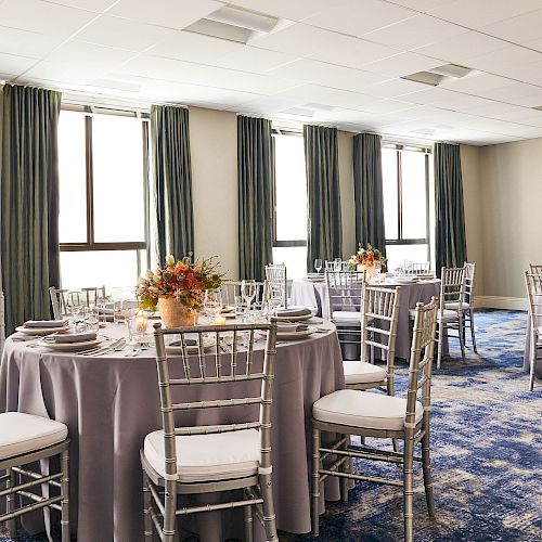 A banquet room with round tables dressed in grey linens, silver chairs, and floral centerpieces, all set on a blue patterned carpet with natural lighting.