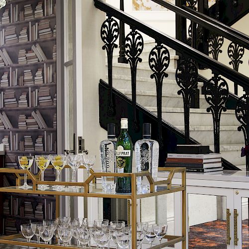 A stylish interior features a stocked bar cart with bottles and glasses, a book-themed wallpaper, and an ornate staircase railing in the background.