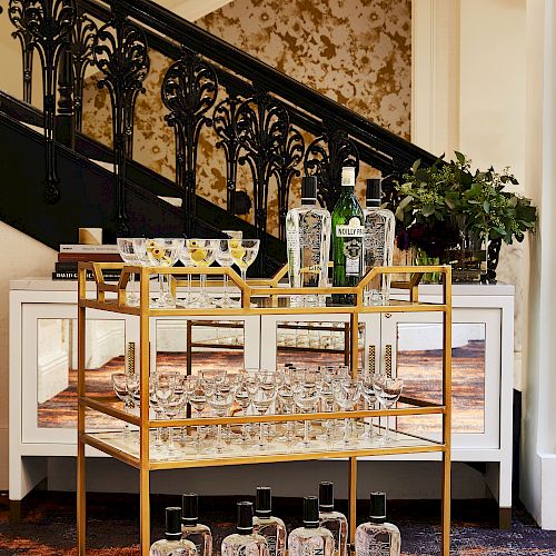 A stylish bar cart stocked with glassware and bottles is positioned in front of an ornate staircase with patterned wallpaper in the background.