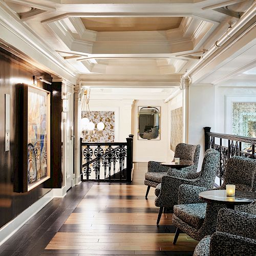 A luxurious hallway features patterned chairs, round tables, artwork, elegant lighting, and a decorative railing along a polished wooden floor.