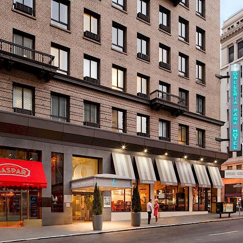 The image shows a street view of a building with shops at the ground level, including a store with a red awning and a green sign for an adjacent hotel.