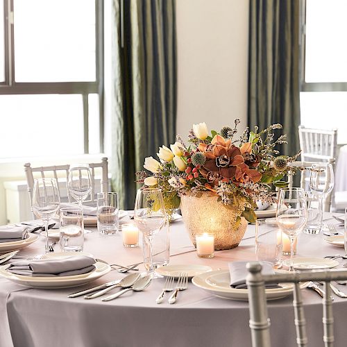 A beautifully set round table with elegant dishware, a floral centerpiece, and candles in a well-lit room with tall windows and draped curtains.