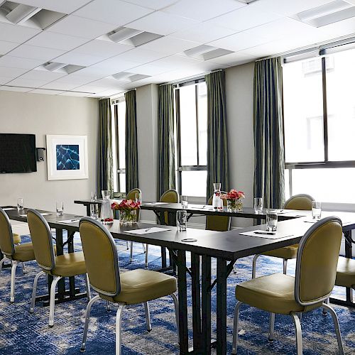 The image shows a conference room with 10 chairs around a U-shaped table, notebooks, pens, water glasses, and flowers, plus large windows and framed art.