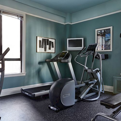 The image shows a small home gym with exercise equipment including dumbbells, a treadmill, an elliptical machine, and a weight bench.