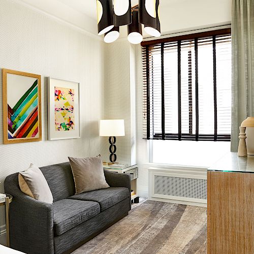 A small living room with a gray sofa, two colorful framed artworks, a side table with flowers, a lamp, a flat-screen TV, and window blinds.