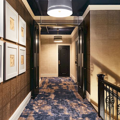 A hallway with framed artwork on the left wall, two doors at the end, patterned carpet, and a railing on the right side under modern lighting fixtures.