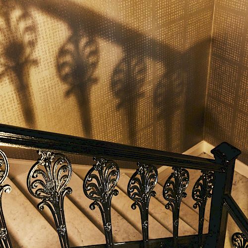 An ornate iron staircase railing casts intricate shadows on a textured wall, leading down a marble staircase.