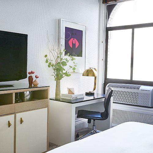 A room with a TV on a stand, a desk with a lamp and chair, a window with an air conditioner, and a bed in the foreground.