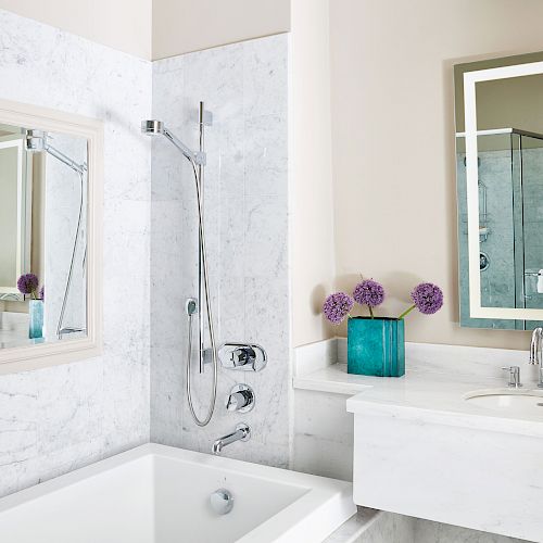 A modern bathroom with a bathtub, vanity with sink, and mirror. There are purple flowers in a turquoise vase on the counter.