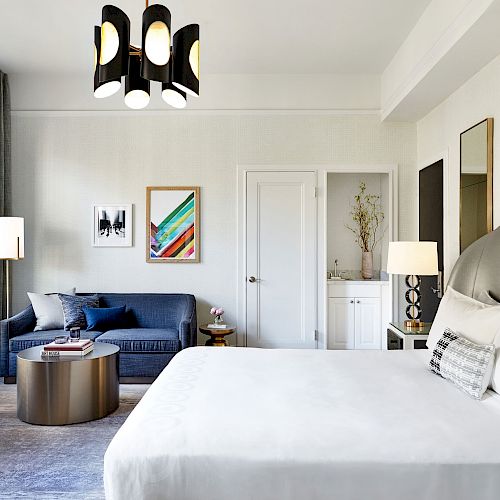 A stylish bedroom with a large bed, a blue sofa, a round coffee table, and modern decor, including a chandelier and framed art on the walls.