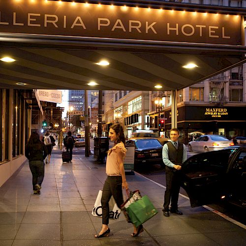 People walk and carry shopping bags near the entrance of Galleria Park Hotel under illuminated signage in a busy city street at dusk.