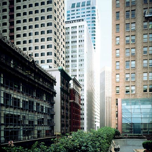 The image shows a cityscape with tall buildings surrounding a narrow, green-lined street or pathway, indicating an urban setting.