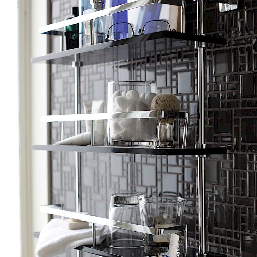 The image shows a wall-mounted bathroom shelf with blue bottles, toiletries, cotton balls, a brush, and a towel, set against a gray patterned wall.