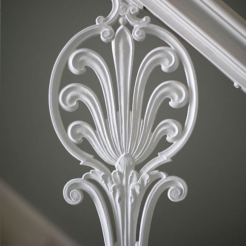 The image shows an intricately designed white wrought iron baluster with a decorative floral pattern, attached to a stair handrail.