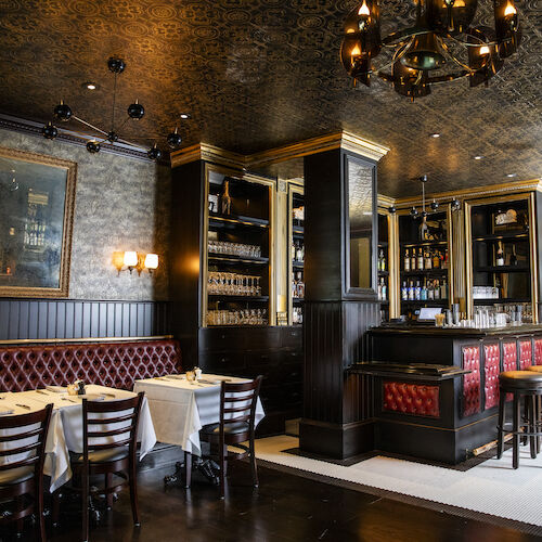 The image shows an elegant restaurant interior with a bar, red leather seating, dark wood accents, hanging lights, and neatly set tables.