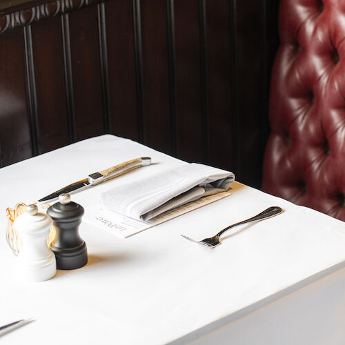 A restaurant table setup with white cloth, napkins, silverware, salt and pepper shakers, and a maroon cushioned booth seat in the background.