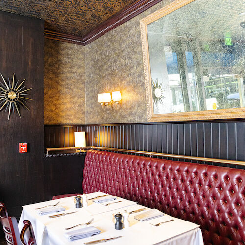 The image shows a cozy restaurant interior with a red tufted booth, a set table, decorative mirror, and wall art.