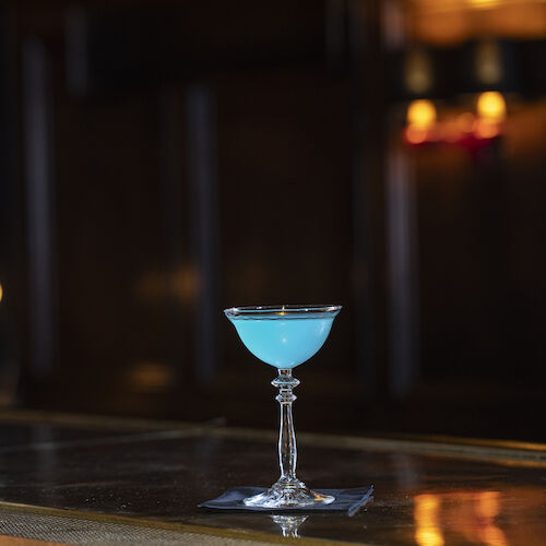 A blue cocktail in a stemmed glass sits on a bar counter with a dimly lit background.