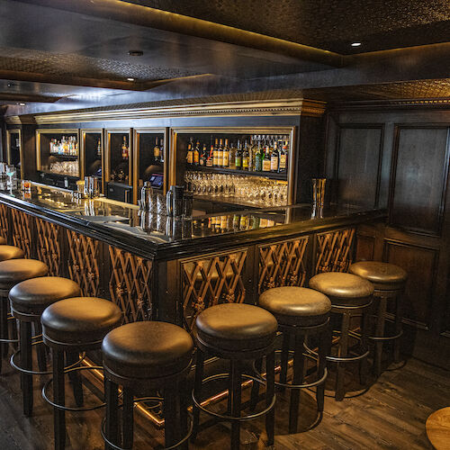 The image shows an empty bar with a long counter, several bar stools, and shelves stocked with various bottles in a dimly lit setting.