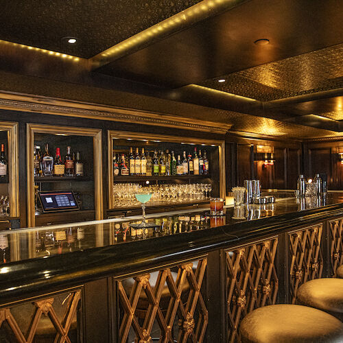 A dimly lit bar with various bottles, glasses, and stools. The atmosphere appears cozy and inviting with elegant wooden decor.