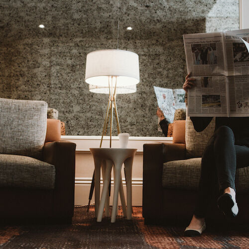 A person is sitting on a chair reading a newspaper with a lamp and side table nearby.