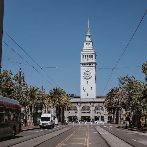 A clock tower stands in the center, surrounded by buildings, palm trees, a bus, and other vehicles on a clear day.
