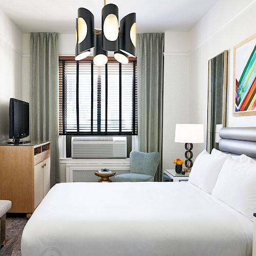 A modern hotel room with a bed, TV, air conditioner, artwork on the walls, a lamp, and a chair by the window. The decor is sleek and contemporary.