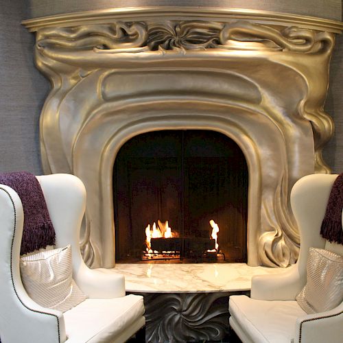 A modern room features a unique, wavy metallic fireplace with a fire burning inside, flanked by two white armchairs with purple throws and pillows.